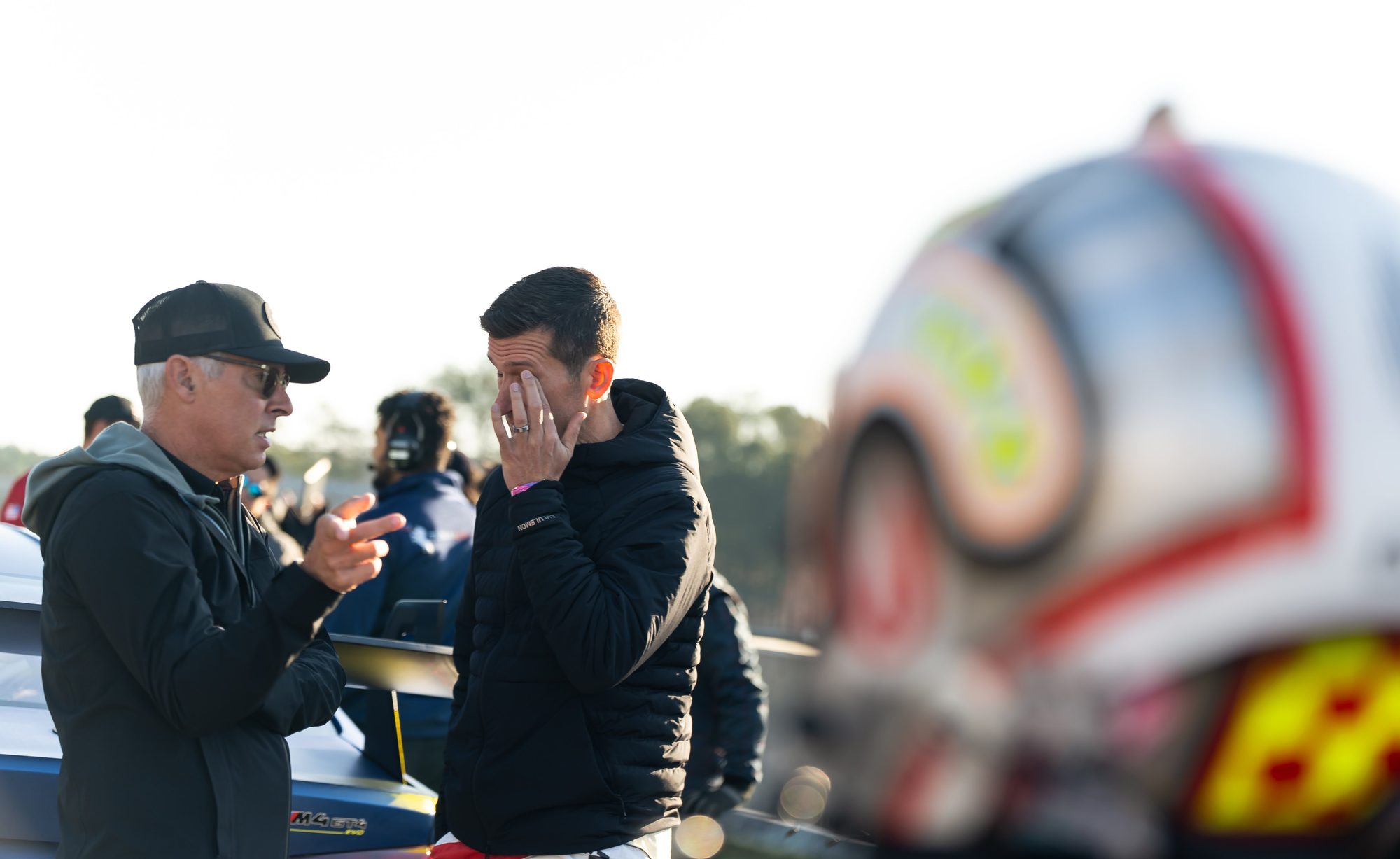Two men talk beside a race car in low sun. One gestures. The other covers his face with a hand, listening.