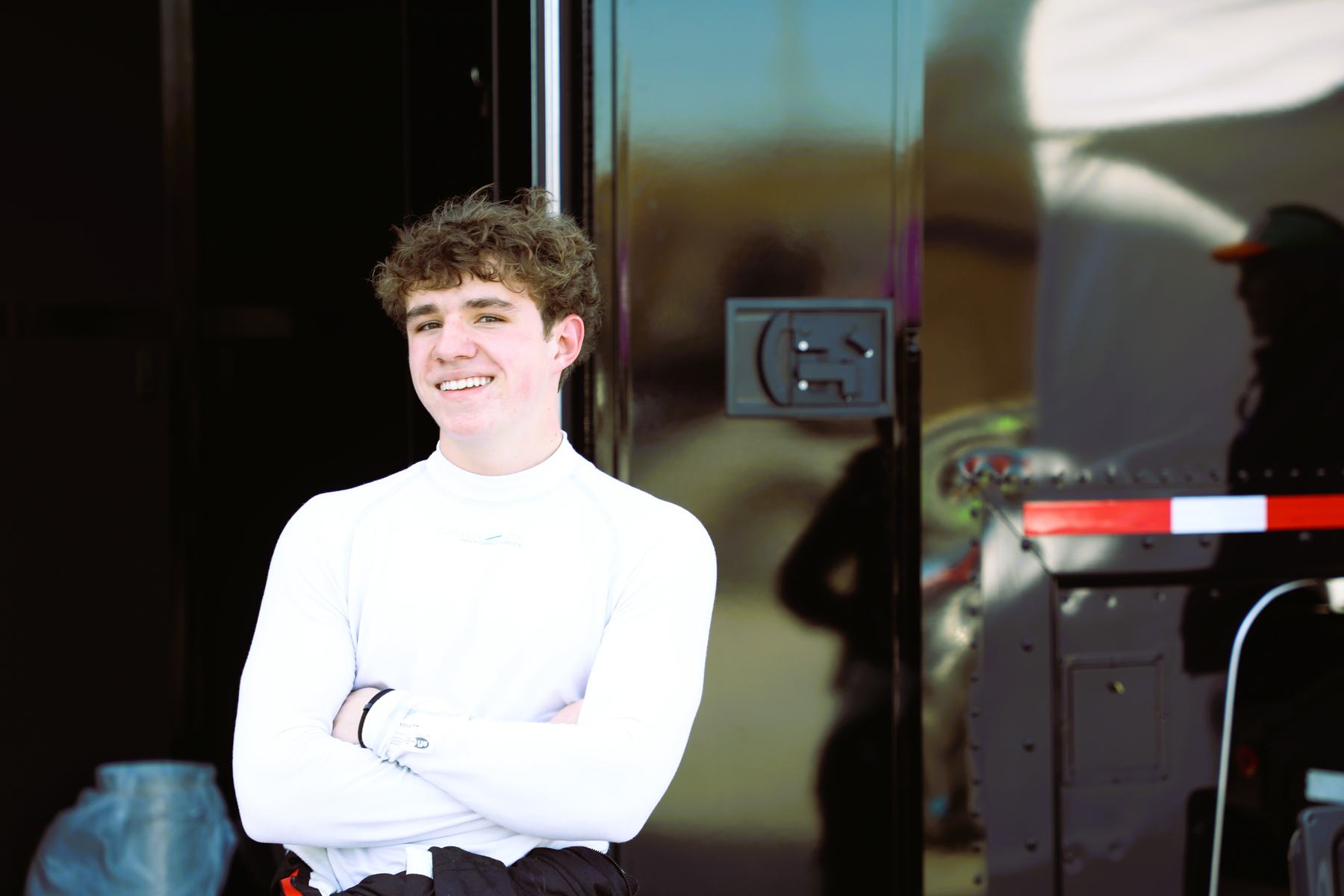 A young driver in a white undershirt smiles with arms crossed by the hauler.