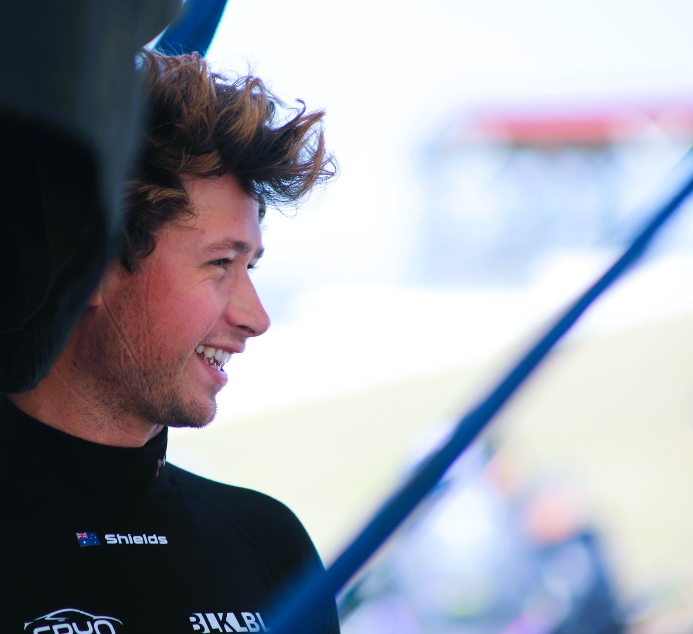 A driver in a black firesuit laughs under the awning, track blurred behind him.