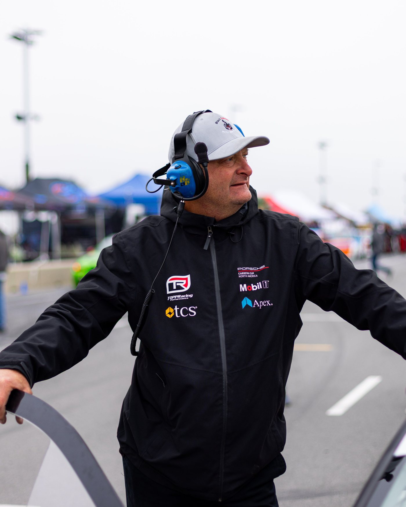 A man in a headset and black jacket holds a car door open in the paddock.