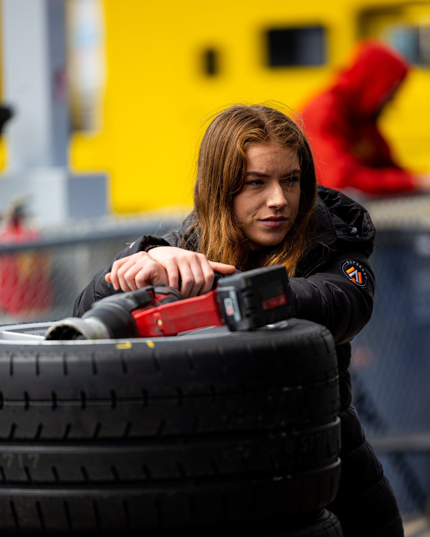 A woman leans on a stack of tires with an impact gun in her hands.