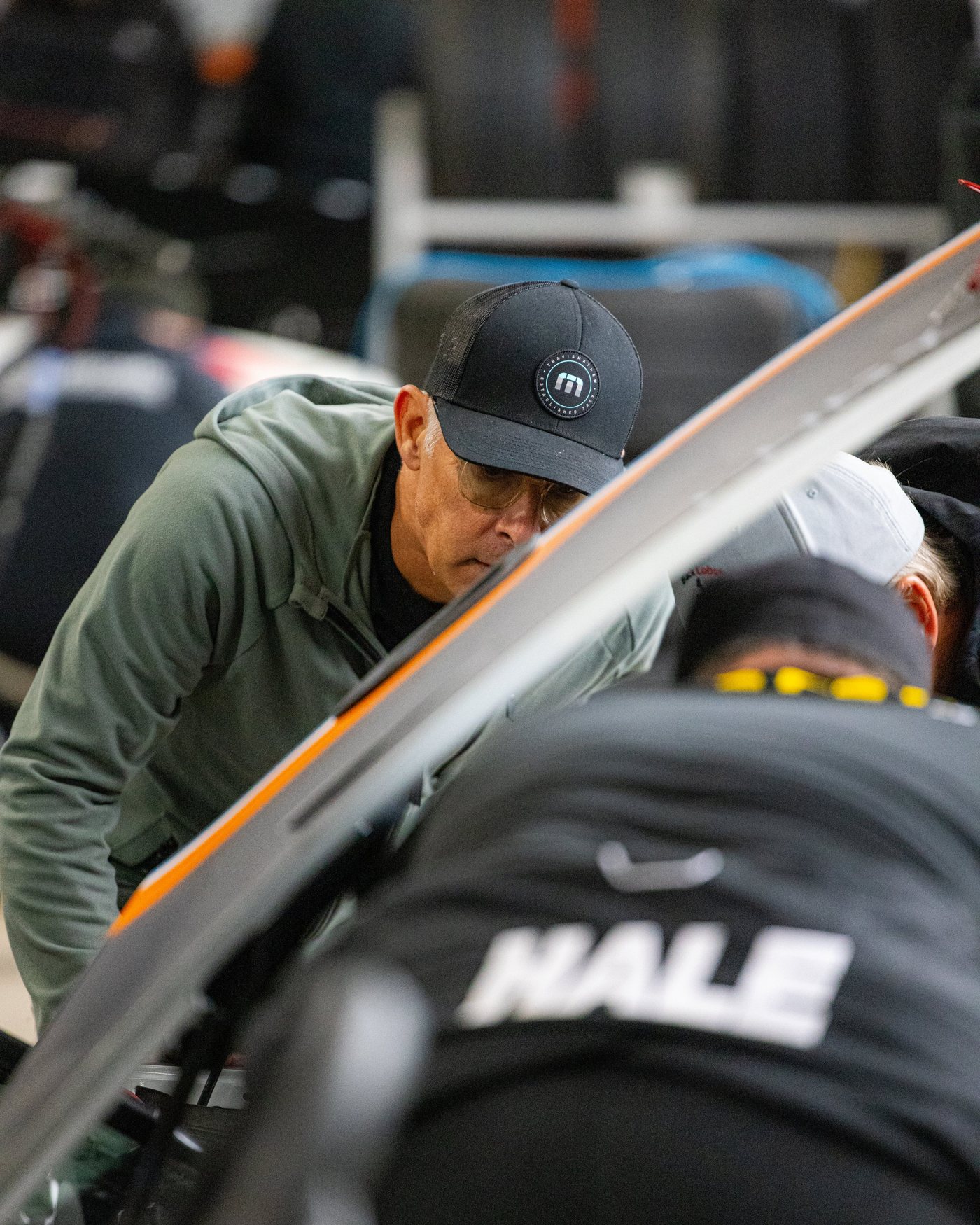 A man in a green hoodie and black cap leans over the car while the crew works underneath.