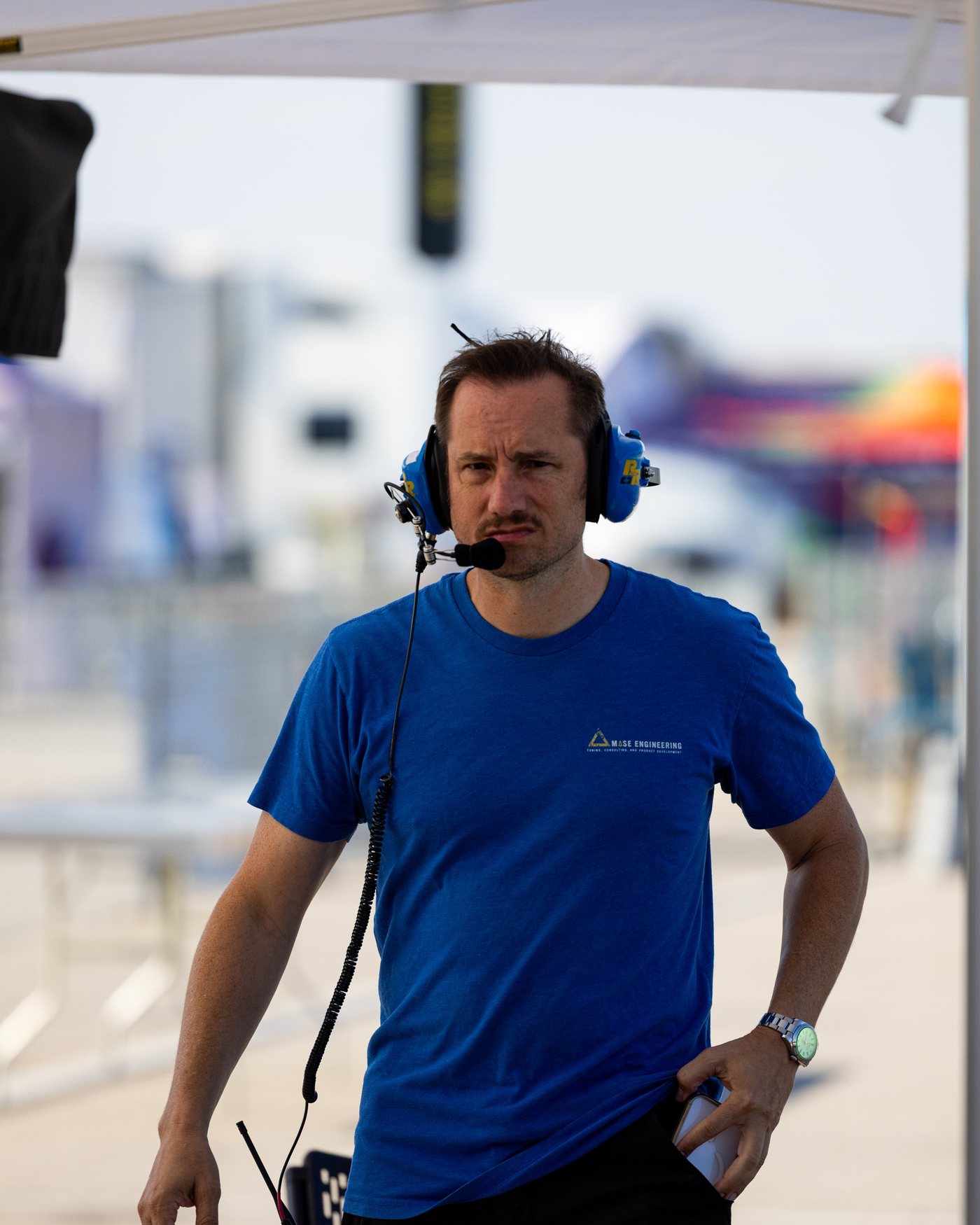 A man in a blue shirt and headset stands under the tent, one hand in his pocket.