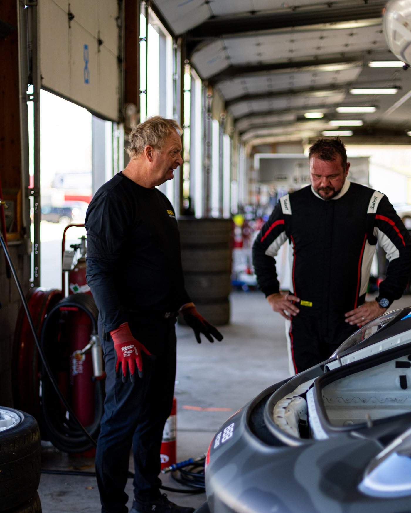 Two men talk beside an open-hood race car in the garage.