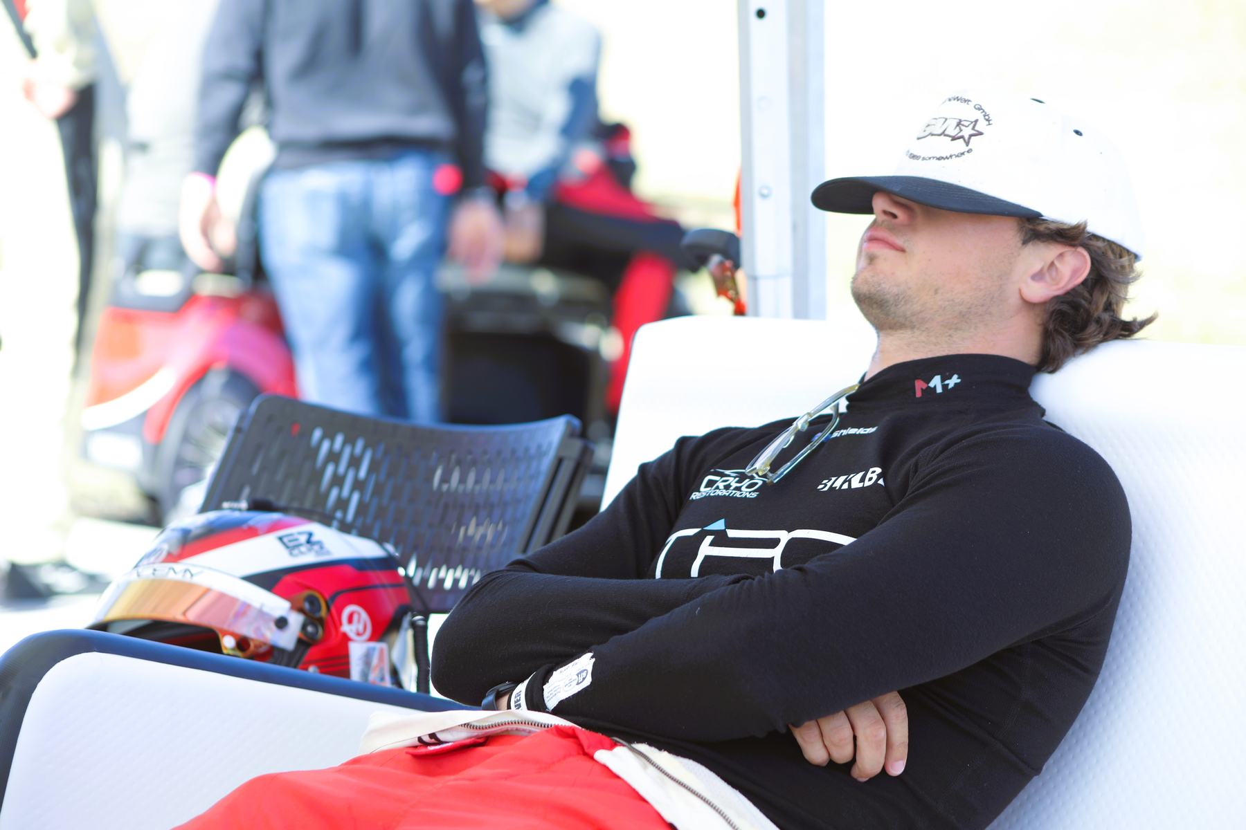 A driver rests in a pit chair, arms crossed, helmet on the ground beside him.